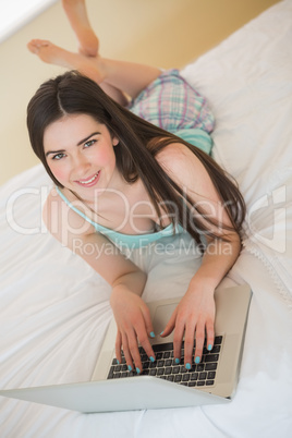 Brunette girl looking at camera and lying on a bed using a lapto