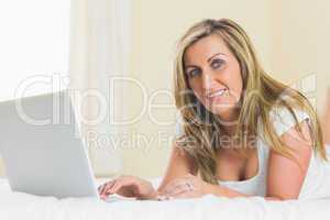 Content woman looking at camera using a laptop lying on a bed