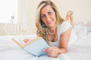 Joyful woman looking at camera reading a book lying on her bed