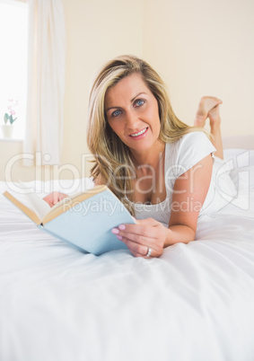 Pleased woman looking at camera reading a book lying on her bed