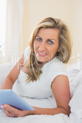 Pleased woman looking at camera using a tablet pc lying on her b