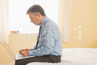 Concentrated man using a laptop sitting on a bed