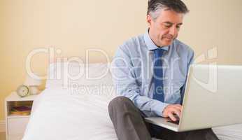 Concentrated man using a laptop sitting on a bed