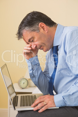 Tired man using a laptop sitting on a bed