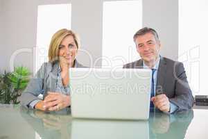 Two smiling business people looking at camera behind a laptop