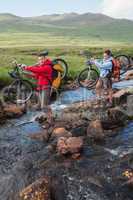 Couple crossing a stream holding their mountain bikes