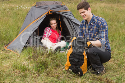 Happy man packing backpack while girlfriend sits in tent