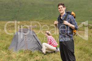 Smiling man carrying backpack while girlfriend is pitching tent