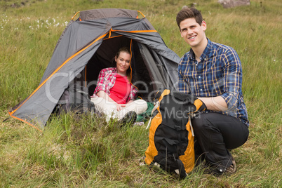 Smiling man packing backpack while girlfriend sits in tent