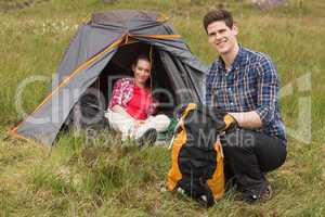 Smiling man packing backpack while girlfriend sits in tent