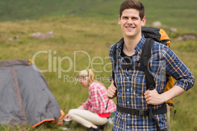 Cheerful man carrying backpack while girlfriend is pitching tent