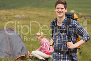 Cheerful man carrying backpack while girlfriend is pitching tent