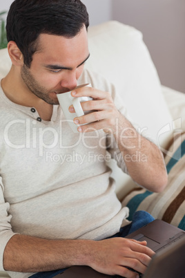 Peaceful attractive man drinking coffee while using his laptop