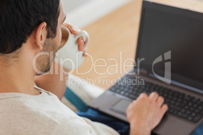 Handsome brown haired man drinking coffee while using his laptop