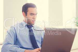 Frowning man biting his eyeglasses and using a laptop