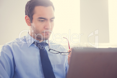 Unsmiling man biting his eyeglasses and using a laptop
