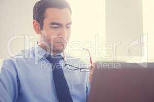 Unsmiling man biting his eyeglasses and using a laptop