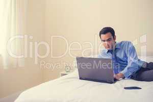 Unsmiling man lying on a bed using a laptop