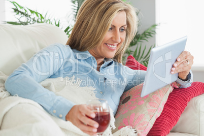Smiling woman lying on a sofa drinking wine and using a tablet p