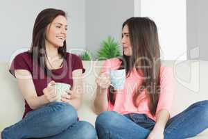 Two girls sitting on a sofa looking each other and drinking a be
