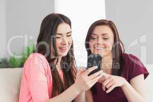 Two smiling girls sitting on a sofa and typing on a mobile phone