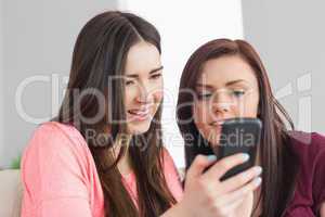 Two happy girls sitting on a sofa and typing on a mobile phone