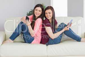 Two smiling girls sitting on a sofa using tablet pc and mobile p