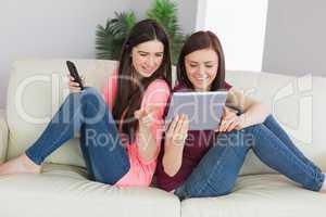 Two happy girls sitting on a sofa using tablet pc and mobile pho