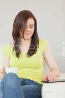 Happy girl sitting on a sofa using a tablet pc and holding a cup