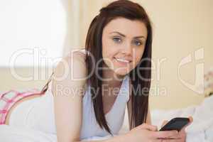 Smiling girl looking at camera using a mobile phone lying on a b