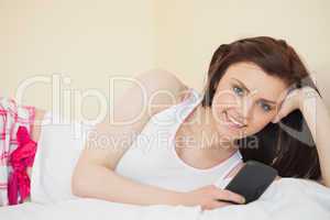 Smiling girl looking at camera using her phone lying on a bed