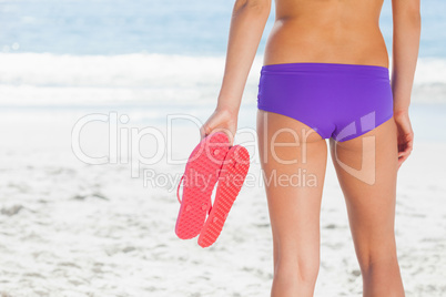 Woman in bikini on beach holding flip flops