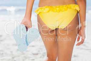 Woman in yellow bikini standing back to camera holding flip flop