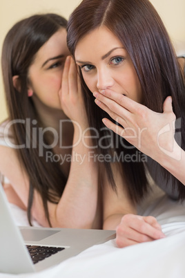 Young girl telling a secret to her friend in front of laptop