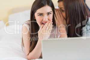 Happy girl telling a secret to her friend in front of laptop