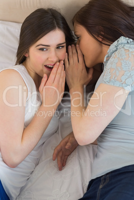 Happy girl telling a secret to her surprised friend lying on bed