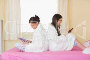 Two friends wearing bathrobes sitting back to back using smartph