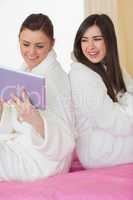 Two smiling friends wearing bathrobes sitting back to back looki