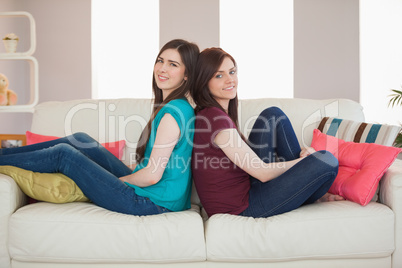 Two smiling friends sitting back to back on the sofa