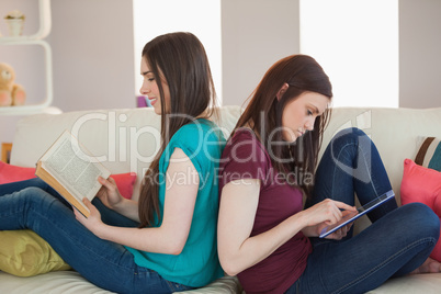 Two happy friends sitting back to back on the sofa reading book