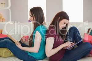 Two happy friends sitting back to back on the sofa reading book