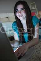 Smiling brunette lying on floor using laptop in the dark looking