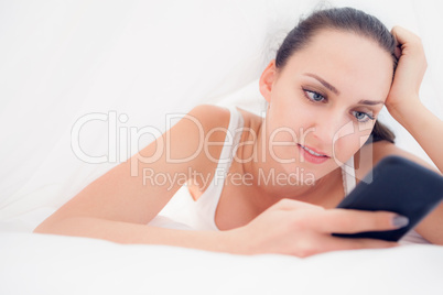 Happy brunette lying under the sheets using her smartphone