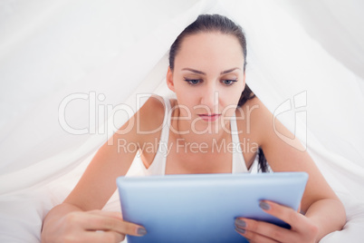 Happy brunette lying under the sheets using her tablet pc