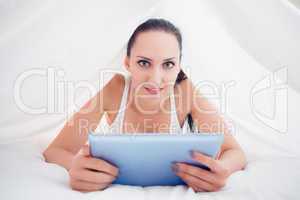Happy brunette lying under the sheets holding her tablet pc