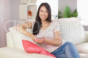 Pretty asian girl using her smartphone on the couch smiling at c