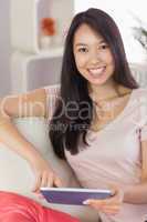 Pretty asian girl using her tablet on the couch smiling at camer