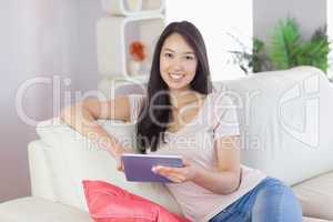 Happy asian girl using her tablet on the couch looking at camera