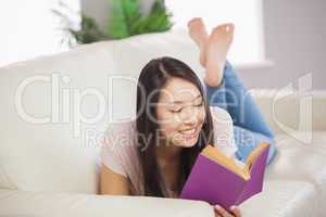 Cheerful asian girl lying on the sofa reading book
