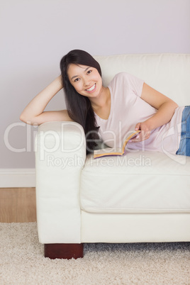 Content asian girl lying on the sofa reading book smiling at cam